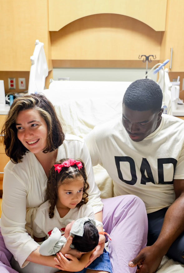 New mom holding baby in her arms surrounded by her husband and first born daughter. Big sister T-shirt. In the hospital on bed with baby. The three of them smiling.